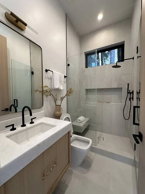 Compact bathroom with a wood vanity, black fixtures and a glass shower below a high window