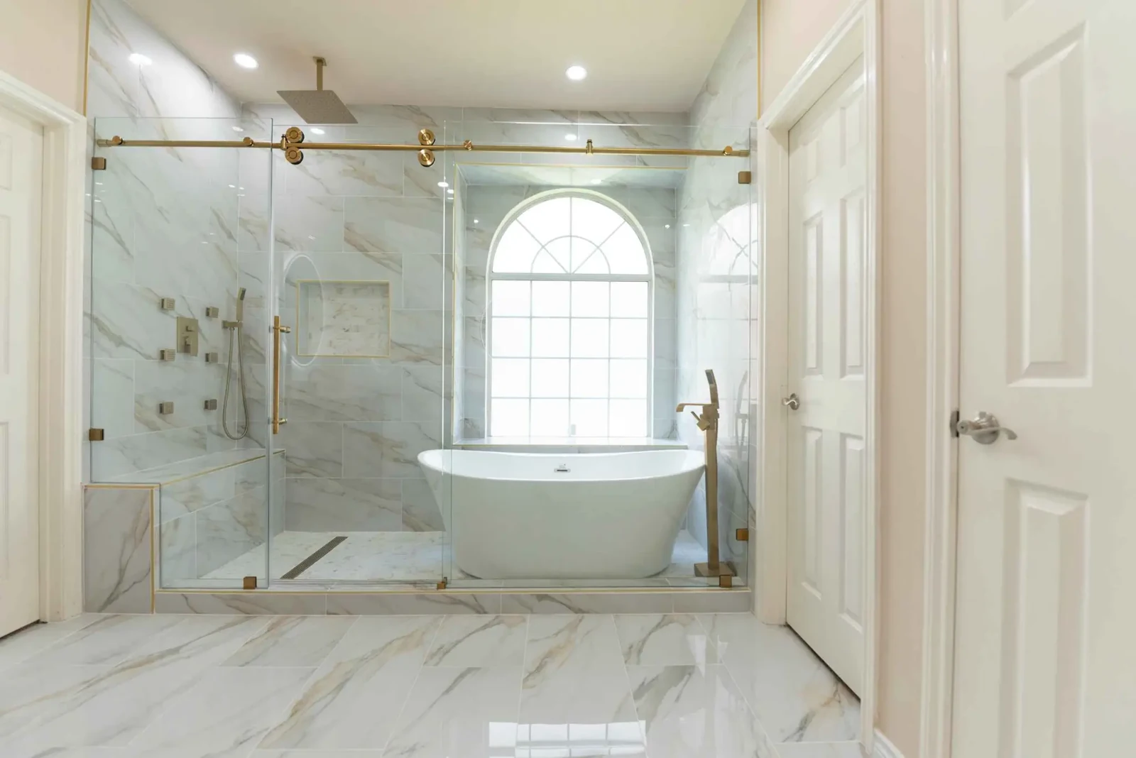 Marble bathroom with brass shower hardware, a glass enclosure and a freestanding tub under an arched window